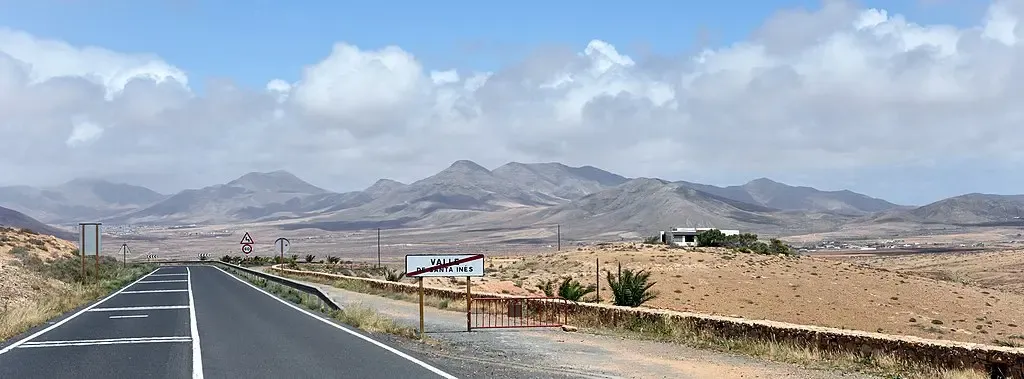 Autocaravana de alquiler circulando por una carretera escénica y vacía de Fuerteventura, rodeada de paisaje volcánico árido y el océano azul al fondo.