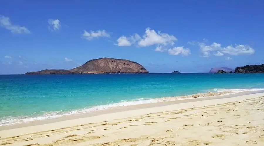 Vista amplia de la Playa de Las Conchas con arena dorada, fuerte oleaje azul y el islote de Montaña Clara al fondo