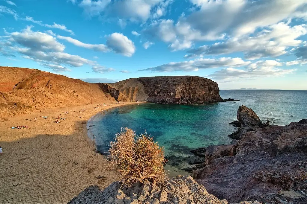 Vista aérea de Playa Papagayo, Lanzarote, destacando su forma perfecta de media luna, arena dorada fina y aguas tranquilas de color turquesa, rodeada por acantilados volcánicos.