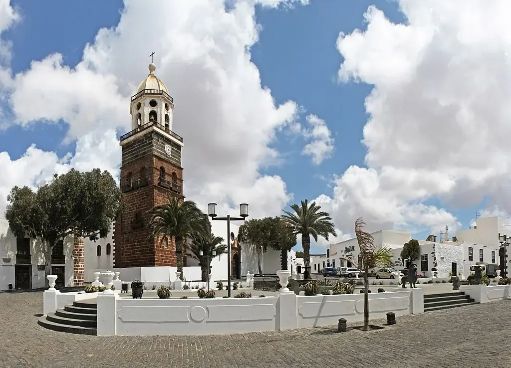 Calle empedrada en el casco histórico de Teguise, Lanzarote, con casas blancas tradicionales de puertas y ventanas verdes y la torre de la iglesia al fondo.